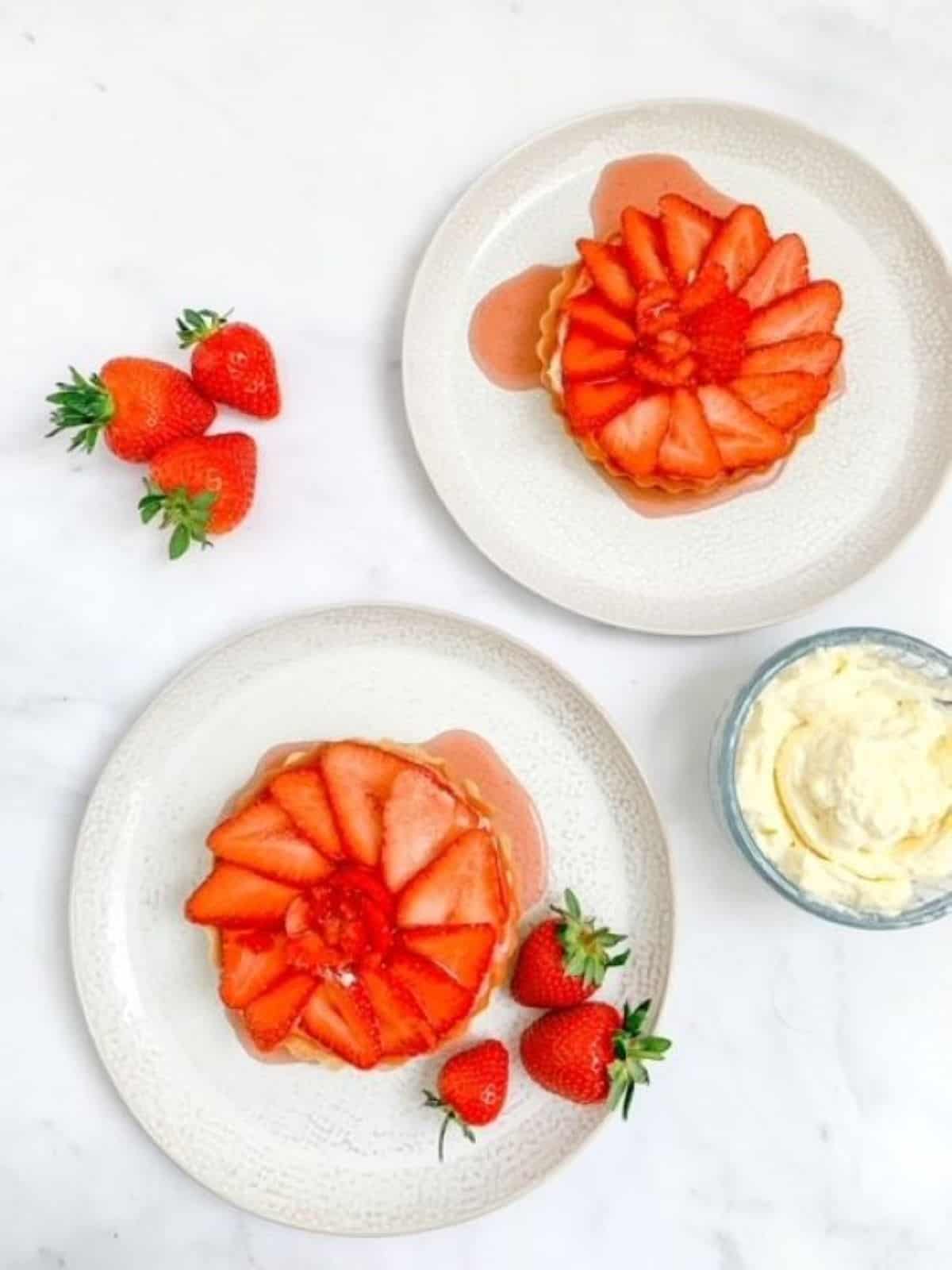 two mini strawberry tartlets on white plates surrounded by fresh strawberries.
