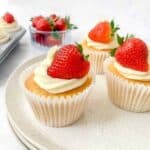 three cupcakes topped with cream and strawberries on a white plate with a bowl of strawberries in the background