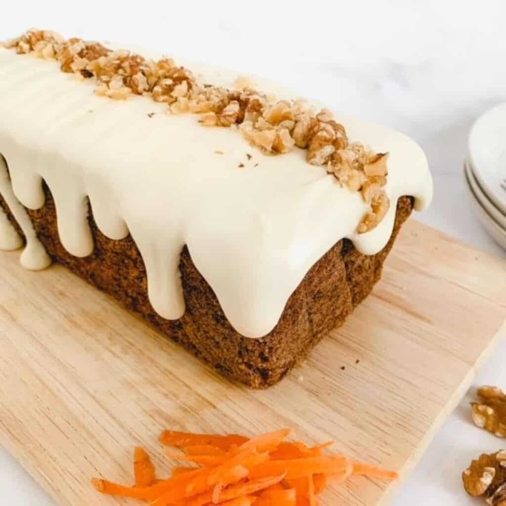 carrot loaf cake with cream cheese frosting topped with chopped walnuts sitting on a chopping board with grated carrot at the side