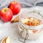 mason jar with overnight oats topped with chopped apple. Two apples and cinnamon sticks in the background