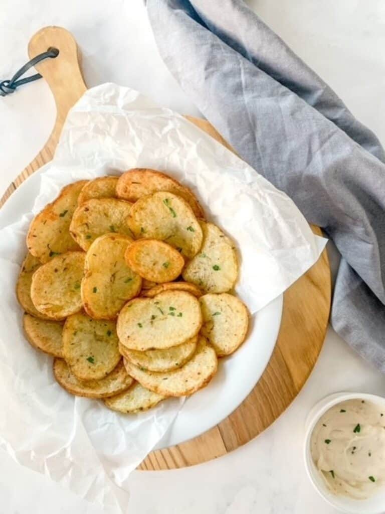 Homemade cottage fries in a white bowl on top of a wooden chopping board beside a napkin and a small pot of garlic dip