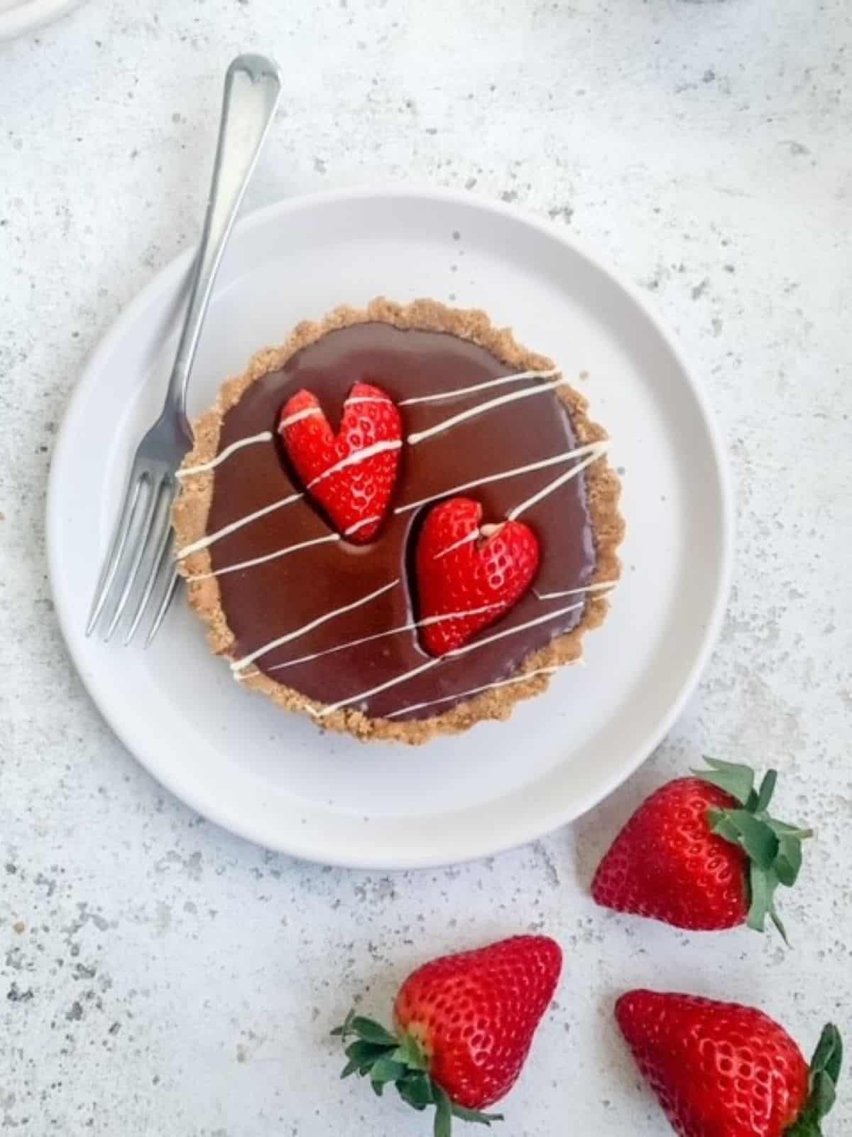 Chocolate ganache tartlet topped with heart shaped strawberries and drizzled with white chocolate with 3 fresh strawberries on the side.