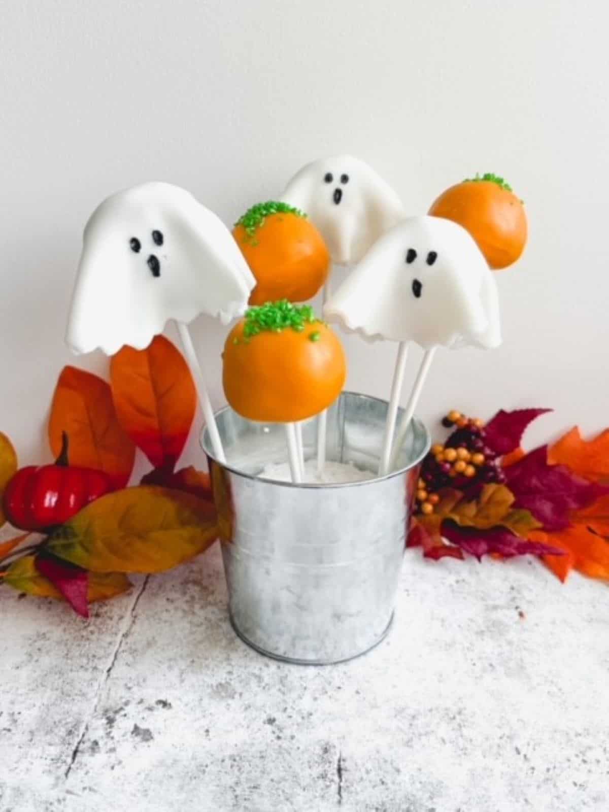 Ghost and pumpkin cake pops in a silver tin surrounded by autumn leaves.