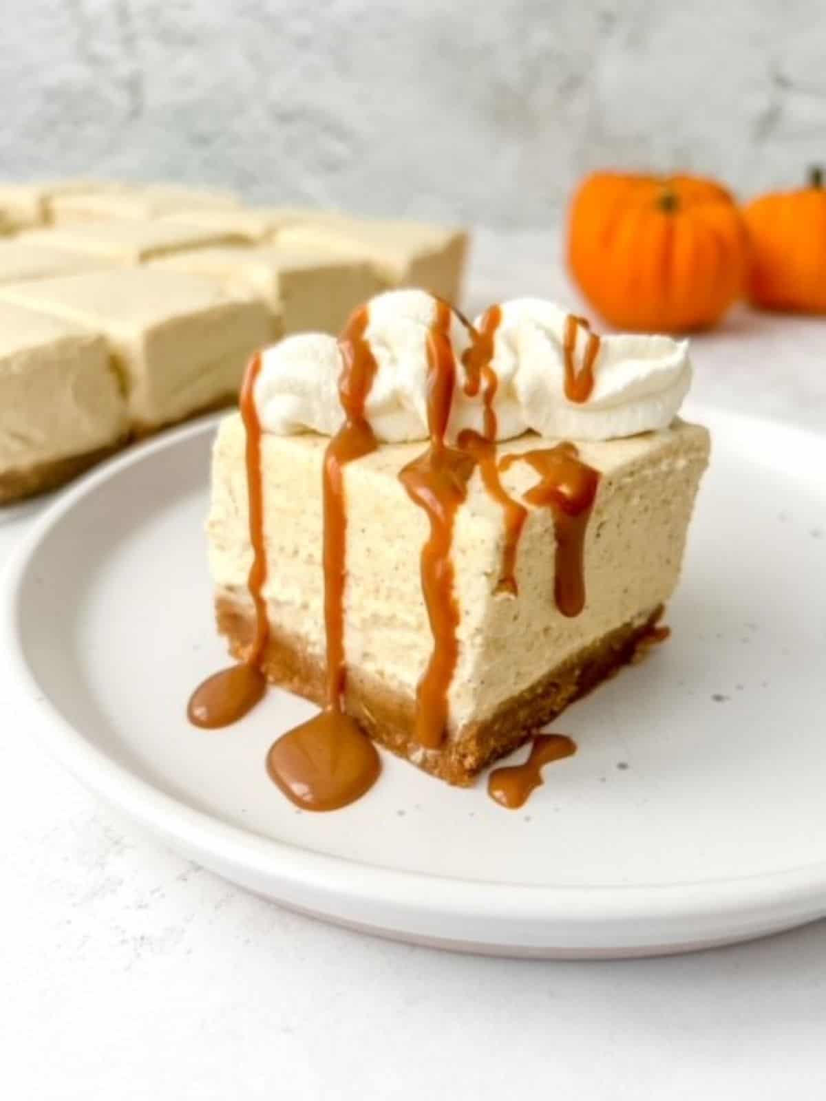 A slice of no bake pumpkin cheesecake bars with cream topping and drizzled in caramel sauce on a white plate.