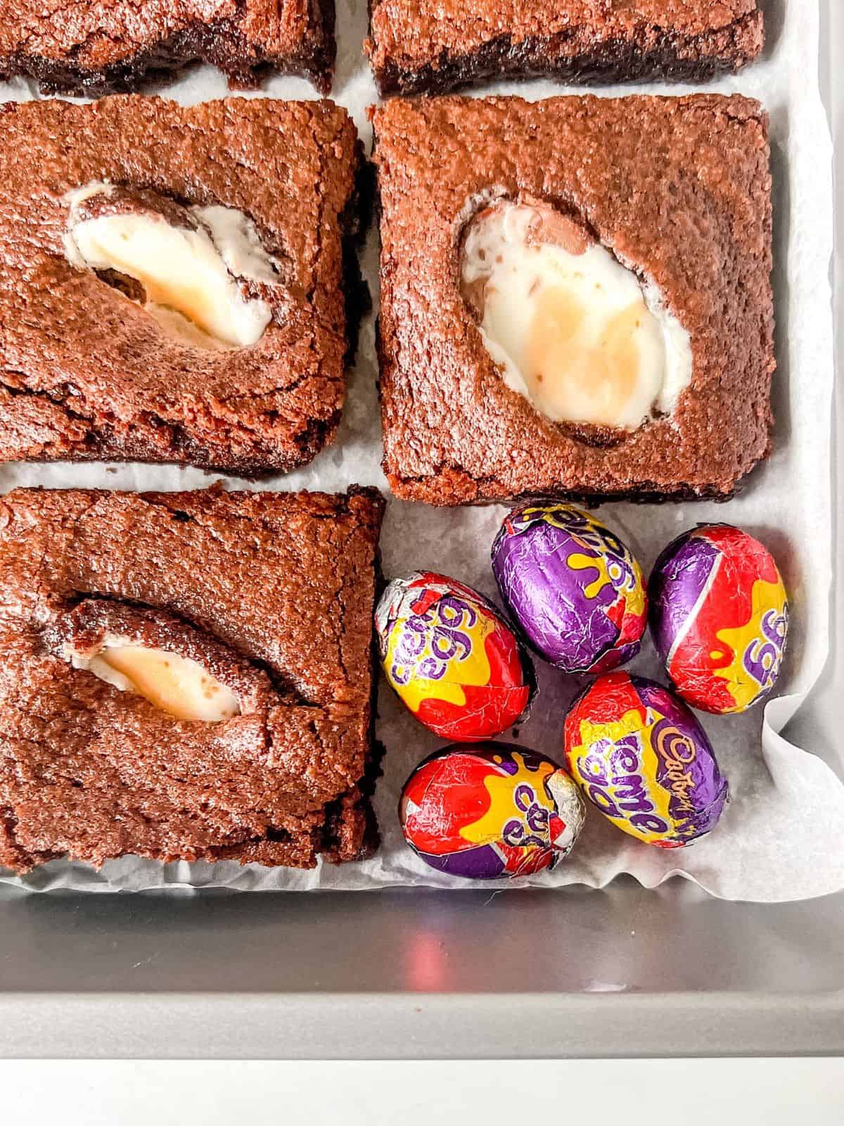 Sliced crème egg brownies in a baking tin with Cadbury creme eggs on the side.
