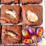 Sliced crème egg brownies in a baking tin with Cadbury creme eggs on the side.
