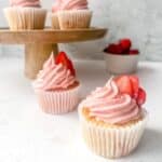 Two gluten free strawberry cupcakes with strawberry buttercream frosting on a white surface with more cupcakes on a cake stand in the background.