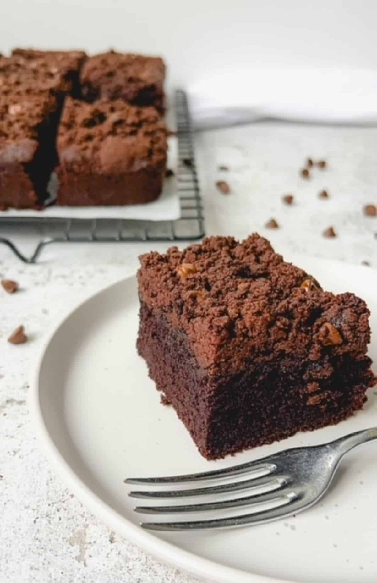 A slice of chocolate sheet cake on a white plate with a fork and the full cake in the background.