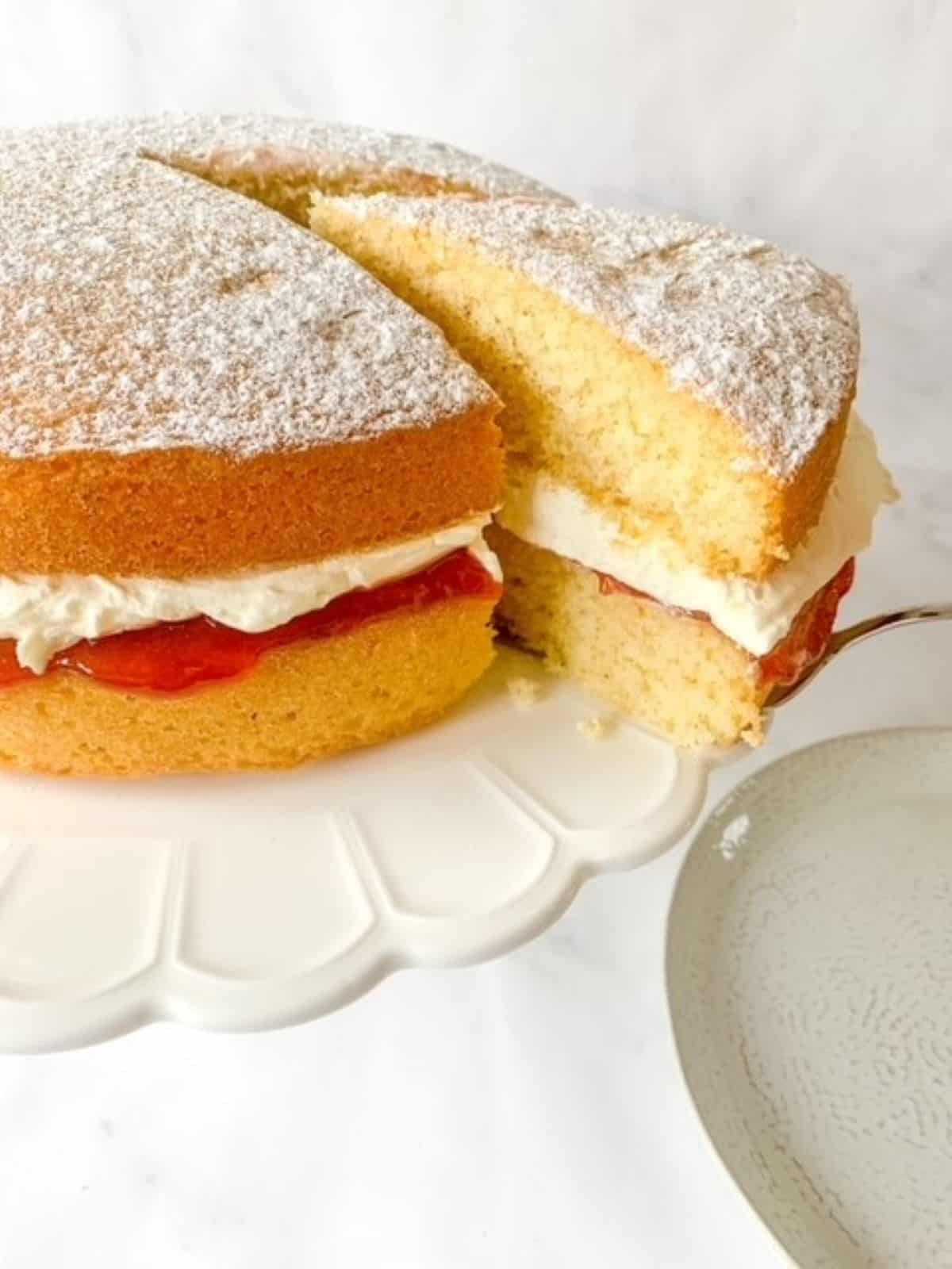 Victoria sponge cake with a slice being removed on a white cake stand.