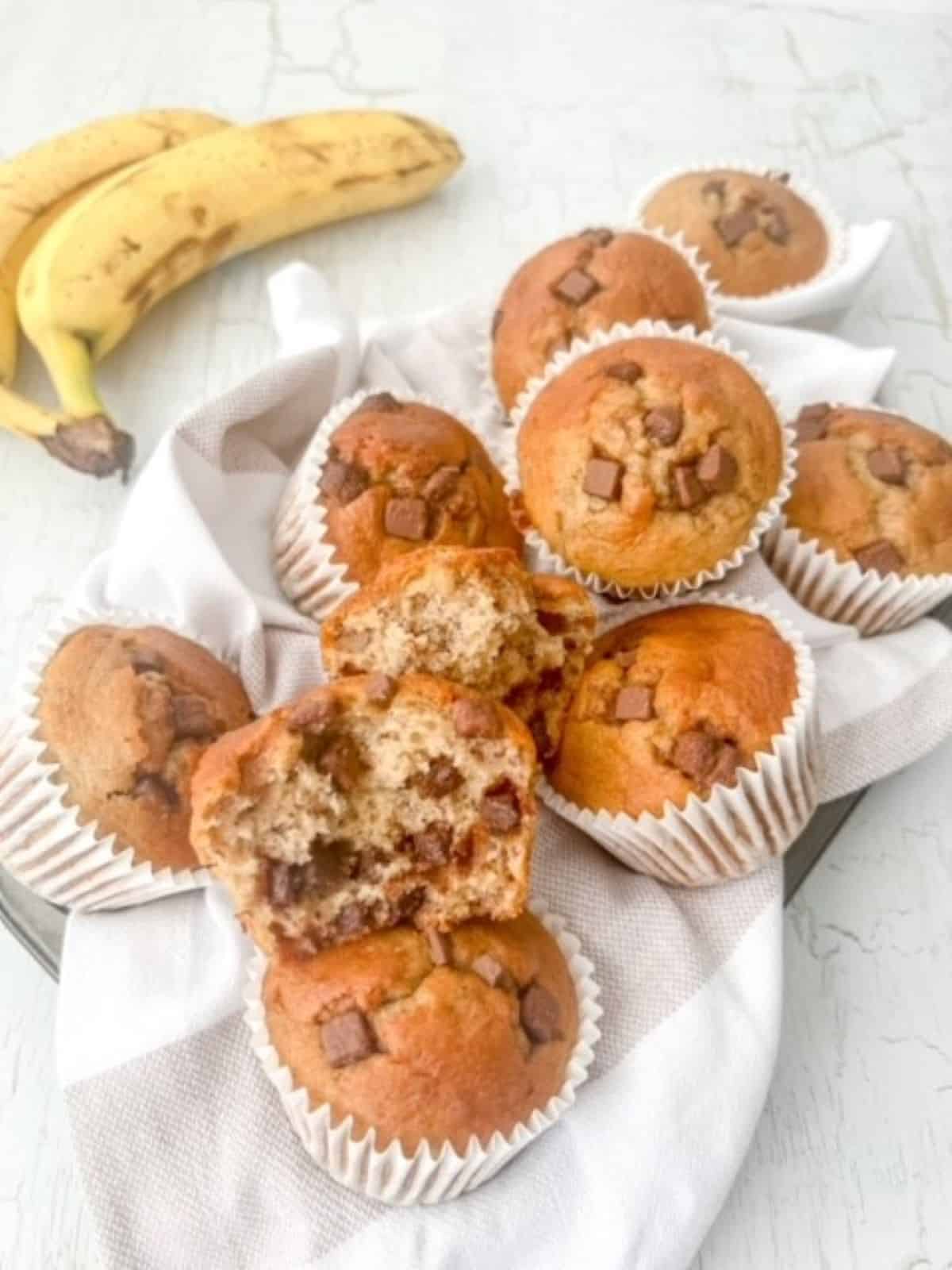 Gluten free chocolate chip banana muffins stacked together on top of a napkin with bananas in the background.