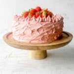 A wooden cake stand with a gluten free strawberry cake topped with fresh strawberries on a white background.