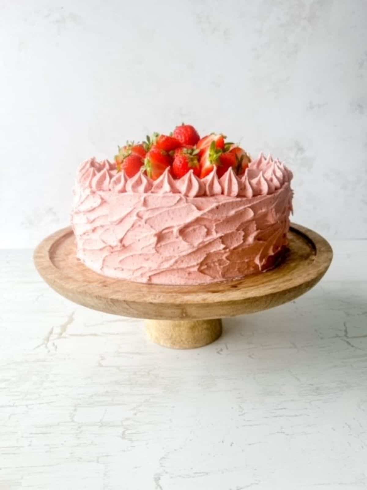 A wooden cake stand with a gluten free strawberry cake topped with fresh strawberries on a white background.