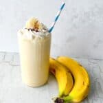 Thick banana milkshake in a tall glass topped with whipped cream beside two ripe bananas.