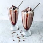 Two chocolate milkshakes in tall glasses topped with whipped cream and chocolate chips.