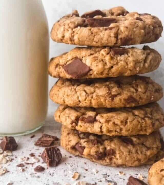 A stack of gluten free oatmeal chocolate chip cookies beside a glass bottle of milk surrounded by chocolate chips and oats.