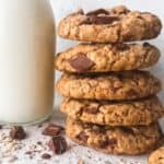 A stack of gluten free oatmeal chocolate chip cookies beside a glass bottle of milk surrounded by chocolate chips and oats.