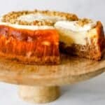 Carrot cake cheesecake on a wooden cake stand with a slice being removed.