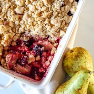 Pear and blackberry crumble in a white dish with pears on the side.