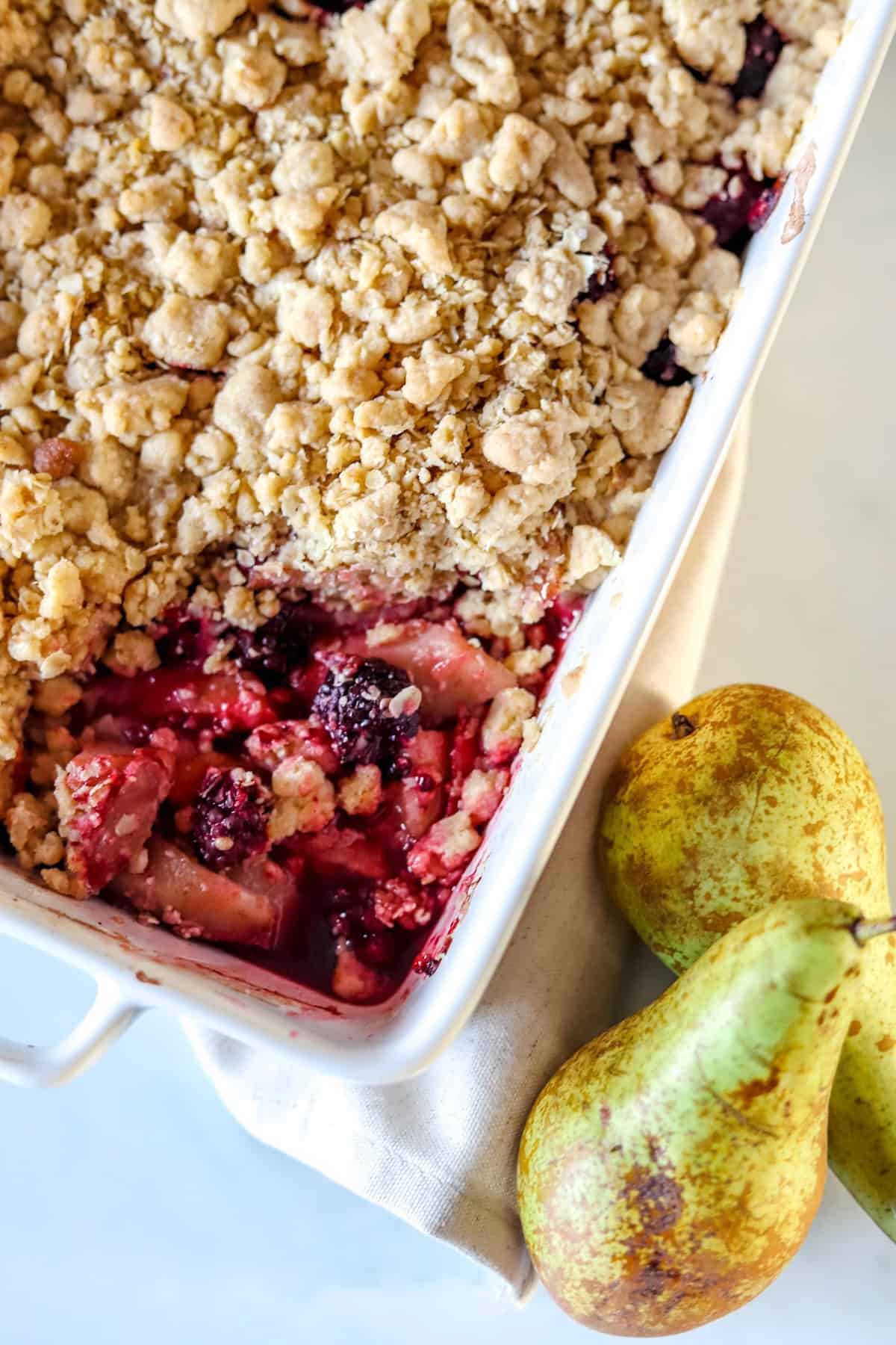 Pear and blackberry crumble in a white dish with pears on the side.