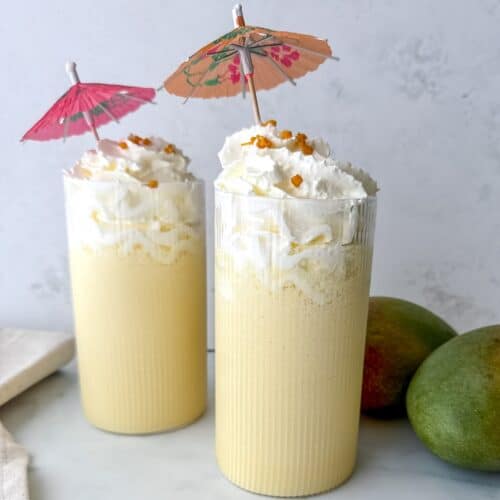 Two tall glasses filled with mango milkshake and topped with whipped cream and a tiny umbrella.