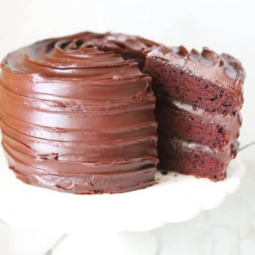 Gluten free chocolate fudge cake on a white cake stand with a slice being removed.